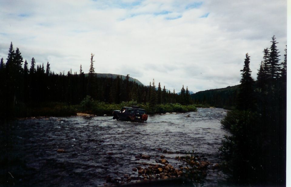 SUV driving through a shallow river with trees and mountains in the background