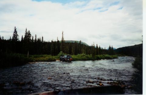 1978 Ford Bronco - A dark SUV navigates a rocky riverbed, surrounded by dense evergreen forests under a cloudy sky.