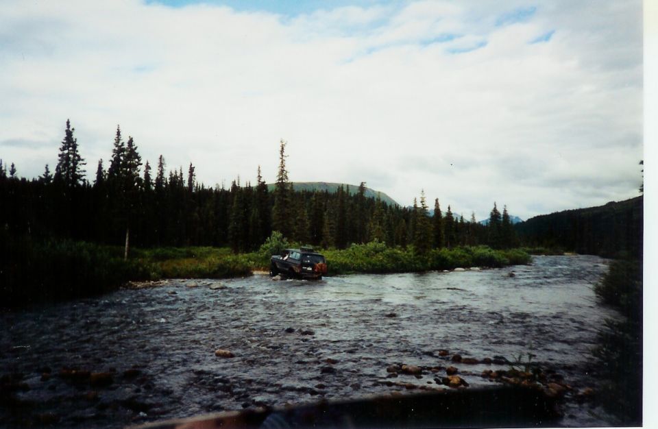 Dark SUV crossing river, rear view. Forested mountains in background