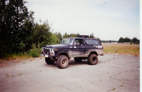 1978 Ford Bronco - A lifted, muddy 1978 Ford Bronco sits parked on an asphalt surface, with trees and a field visible in the distance.