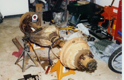 1978 Ford Bronco - Front axle assembly with drum brakes supported by jack stands in a cluttered garage setting.