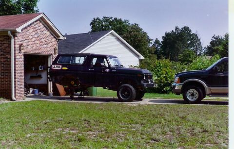 1978 Ford Bronco - A black Ford Bronco is elevated on jack stands in front of a brick garage, with another SUV parked to its right.