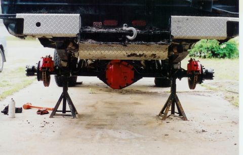 1978 Ford Bronco - The rear axle and suspension of a vehicle are elevated on jack stands, revealing red brake calipers and diamond plate bumper guards.