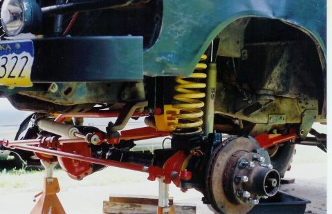 1978 Ford Bronco - Close-up of a green vehicle's front suspension, a yellow coilover shock and red control arms, with an Alaska license plate visible.