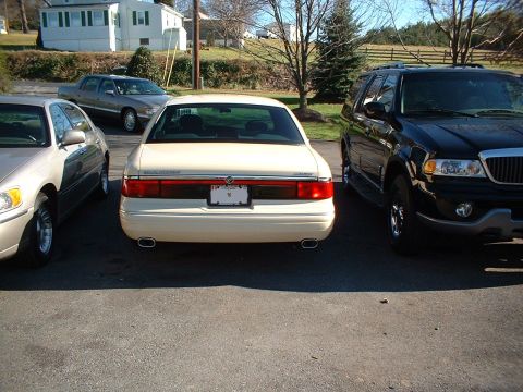 1996 Mercury Grand Marquis - The rear of a cream-colored sedan is visible, parked between a light-colored sedan and a black SUV.
