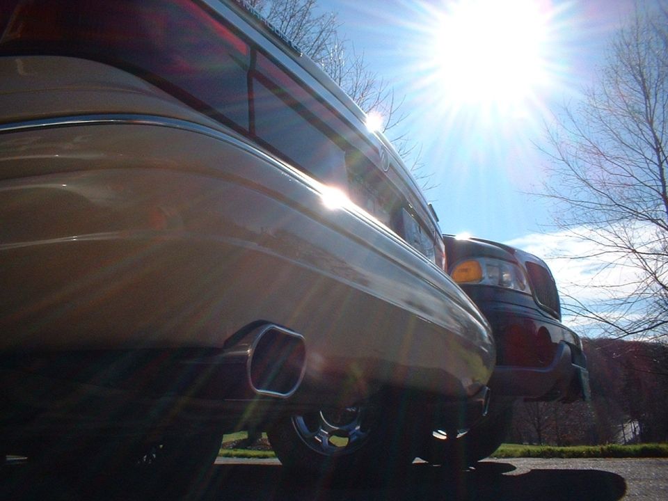 Low-angle rear shot of gold sedan with chrome exhaust tip, sun flare, dark vehicle beside it