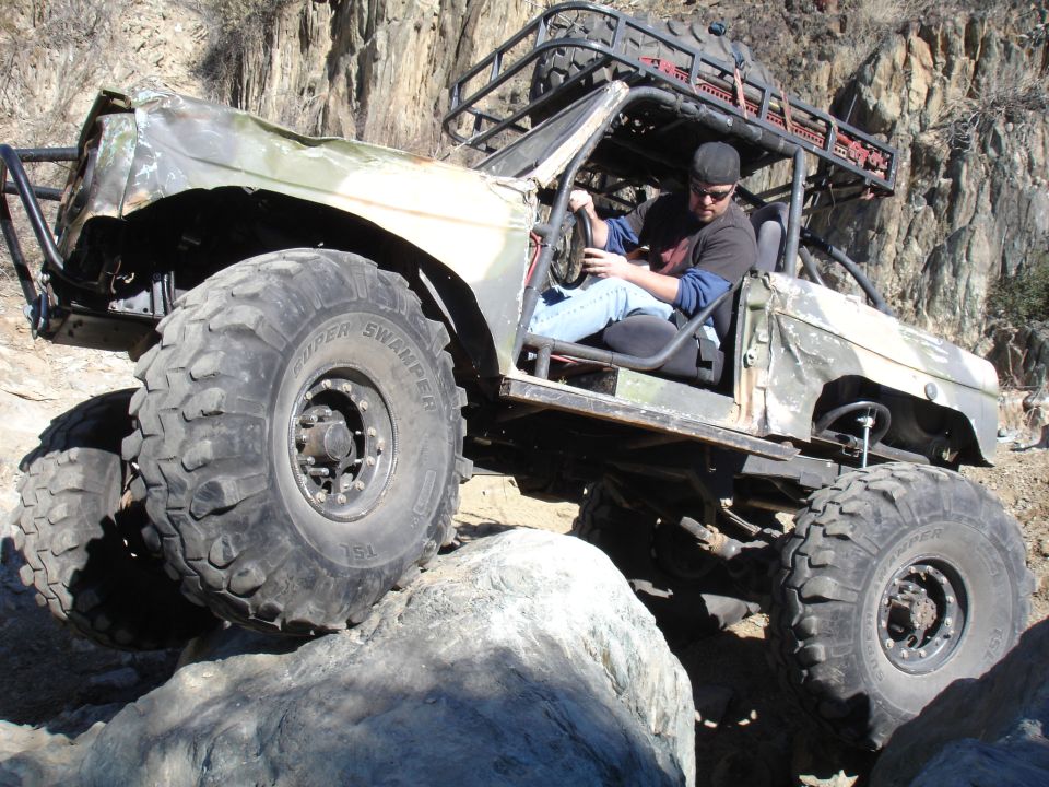 Modified off-road vehicle with large tires climbing over rocks. Driver is visible inside