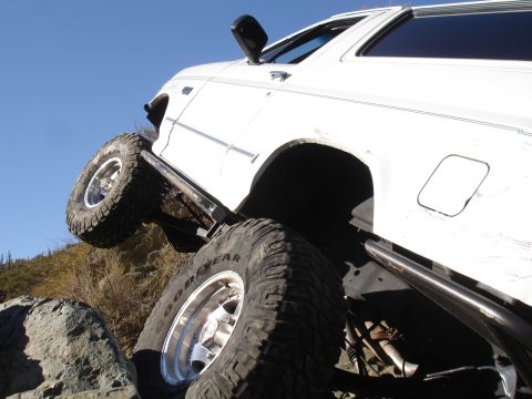 1984 Ford Bronco - Low-angle side view of a lifted white full-size SUV climbing over rock on a desert trail against a deep blue sky, with the rear tire flexed…
