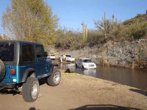 1984 Ford Bronco - Several trucks navigate a shallow stream in a desert environment, with a Jeep parked nearby.