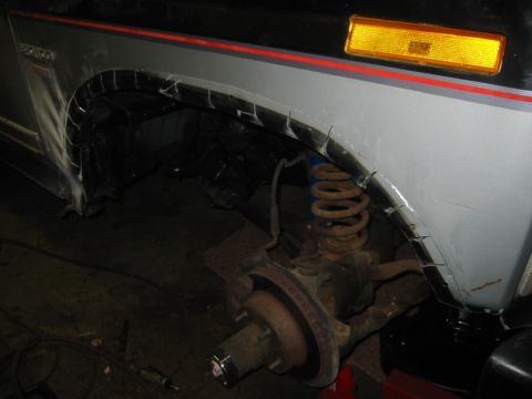 1986 Ford Bronco - The front wheel well of a vehicle shows cut fenders and exposed suspension components, with a gray body and red stripe detail.