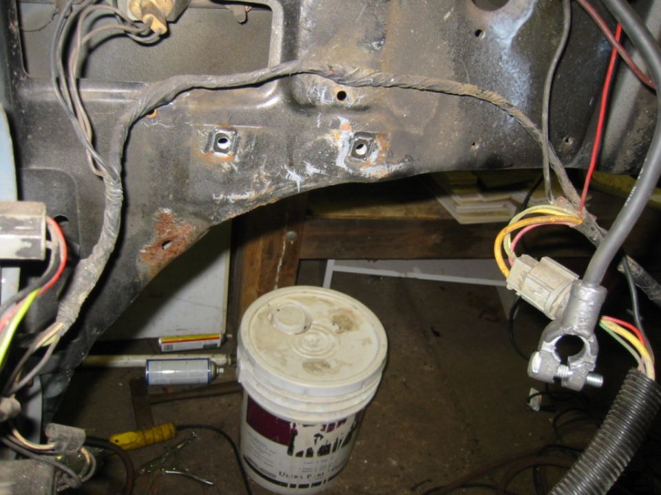 Engine bay wiring harness and frame section with rust spots. Bucket and spray can visible below