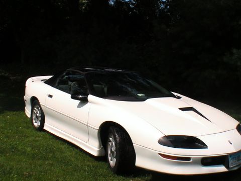 1996 Chevrolet Camaro - A white convertible sports car with a black top and hood scoop is parked on a grassy area with trees behind it.