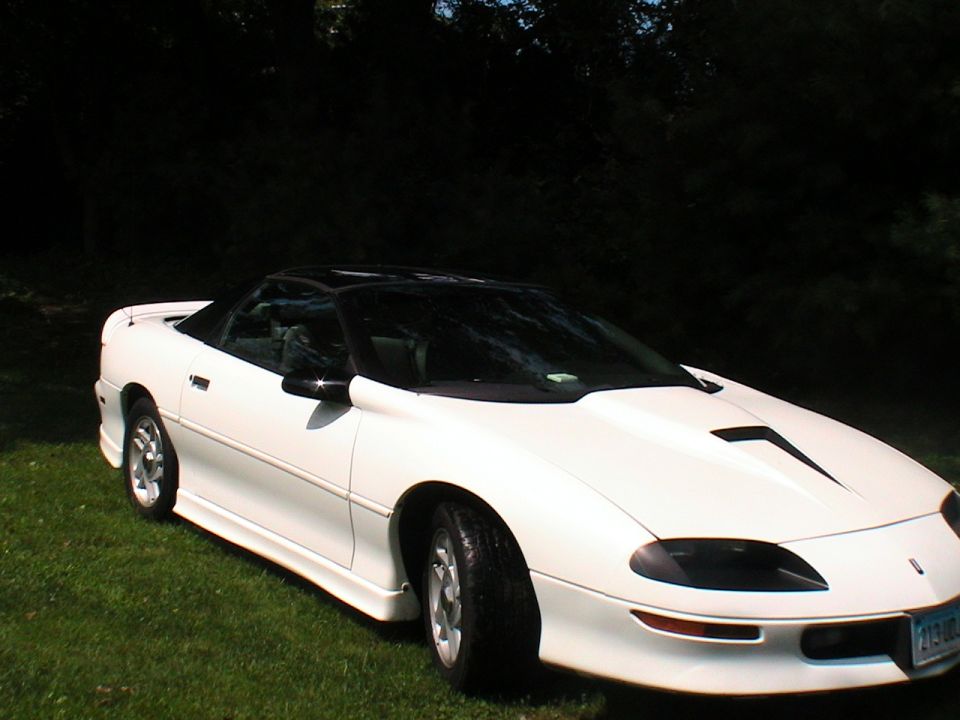 White convertible sports car parked on grass with trees in background. Has a black top and hood scoop