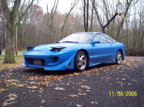 1995 Ford Probe - A blue sports coupe with a custom body kit and pop-up headlights is parked outside in a wooded area.