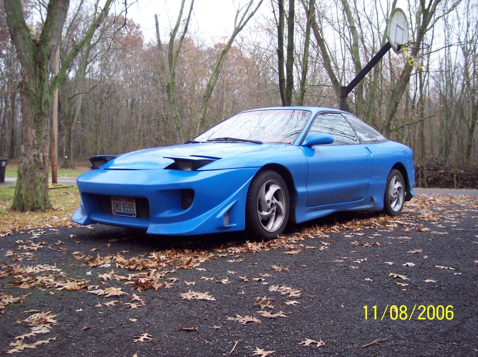 Blue sports coupe with aftermarket body kit and pop-up headlights parked on a driveway with fallen leaves