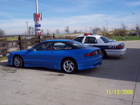 1995 Ford Probe - A blue coupe with an aftermarket body kit is parked next to a police car, with a gas price sign in the background.