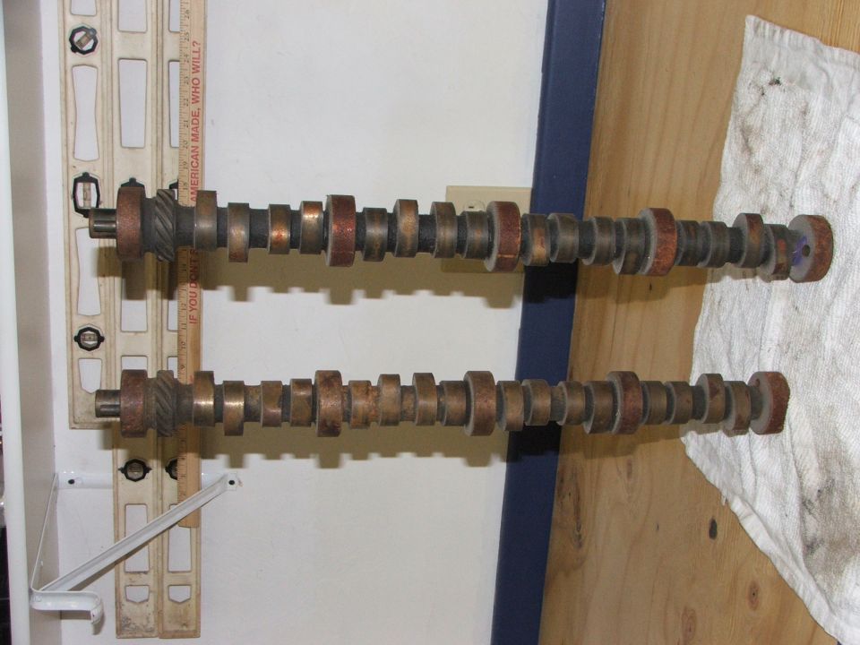 Two camshafts with multiple lobes, one above the other, resting on a wooden surface