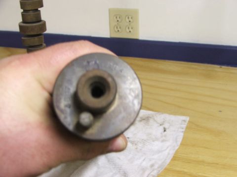 1965 Ford Mustang - A hand holds a metal automotive part, possibly a fuel pump or filter housing, against a wooden surface.