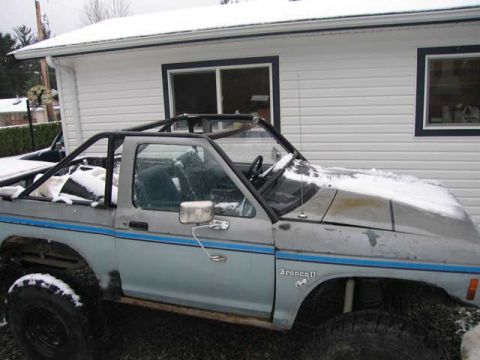 1984 Ford Bronco II - A 1984 Ford Bronco II with a roll cage sits outside a building, partially covered in snow.