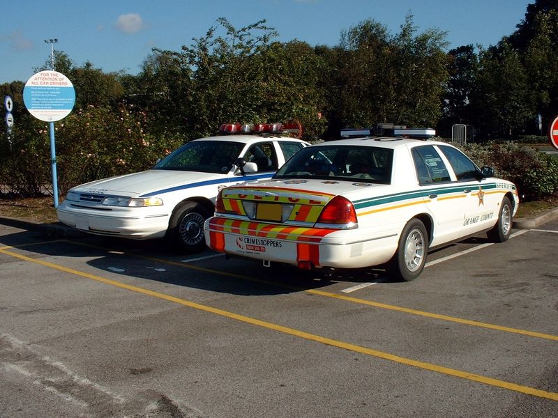 Rear view of white Orange County Sheriff sedan with chevron panel, parked next to another white police car.