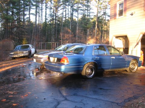 1991 Ford LTD Crown Victoria - A blue sedan is parked next to another car in a driveway with a house and trees in the background.