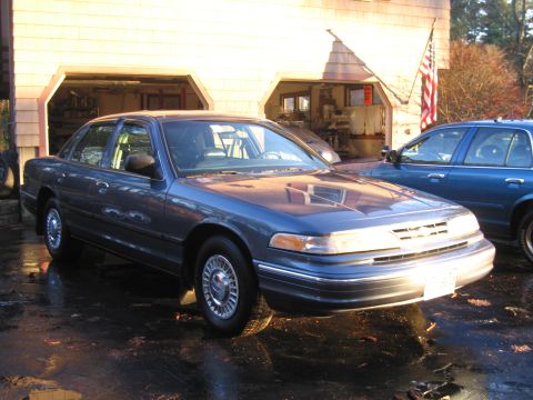 1991 Ford LTD Crown Victoria - A blue Ford Crown Victoria is parked outside a garage, with another blue vehicle visible behind it.