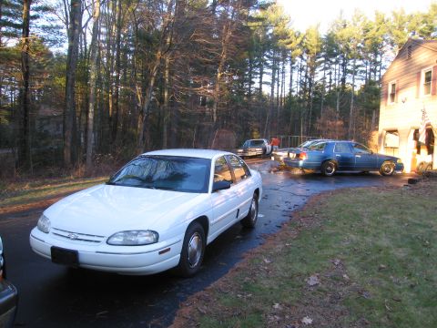 1991 Ford LTD Crown Victoria - A white sedan is parked in a driveway alongside other vehicles, with a wooded area and house visible.