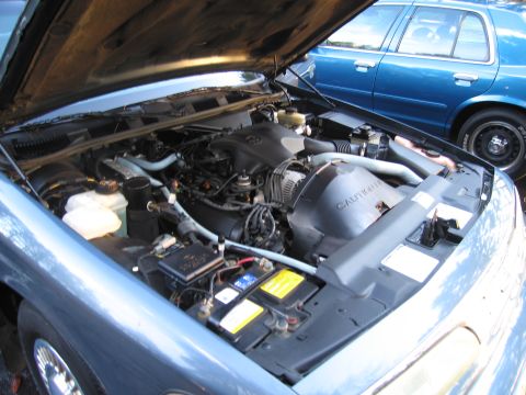 1991 Ford LTD Crown Victoria - Engine bay of a 1991 Ford Crown Victoria LTD, showing the engine and battery. A second blue vehicle is parked behind.