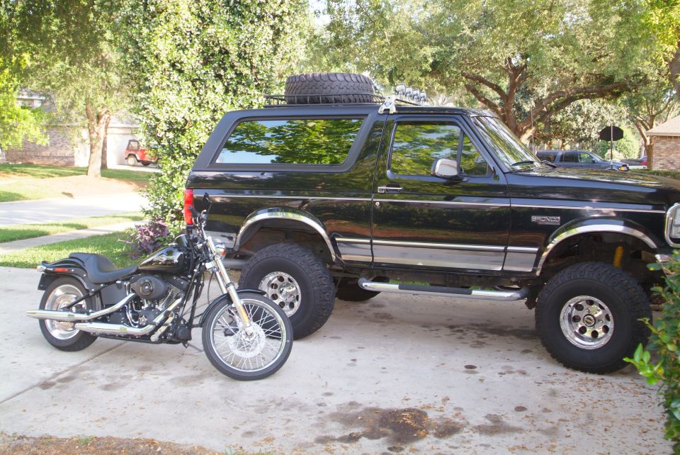 Lifted black full-size Ford Bronco with roof-rack spare on a driveway, black Harley Softail parked alongside