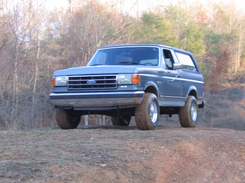 1989 Ford Bronco - A blue Ford Bronco is parked on a dirt incline, with a wooded area behind it.
