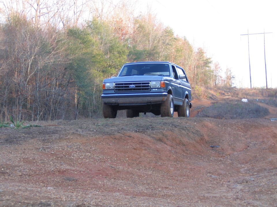 Blue Ford Bronco driving on a dirt hill, front 3/4 view. The vehicle has a blue grille and chrome bumper