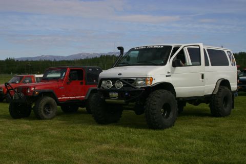 2003 Jeep TJ - Two modified off-road vehicles, a red Jeep Wrangler and a white Ford E-Series van, parked on a grassy field.