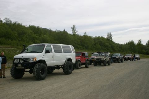 2003 Jeep TJ - A group of off-road vehicles, including a van and several SUVs, are parked on a gravel road with a wooded hillside in the background.