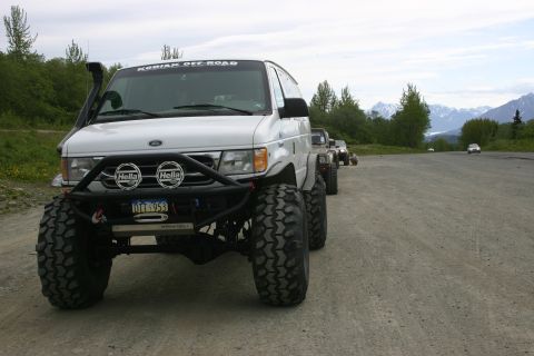 2003 Jeep TJ - Several modified vehicles are parked in a line on a gravel road with mountains in the background.