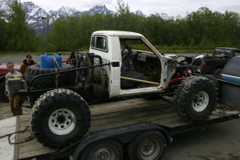 2003 Jeep TJ - A heavily modified pickup truck with oversized tires is being transported on a trailer, with snowy mountains visible in the distance.
