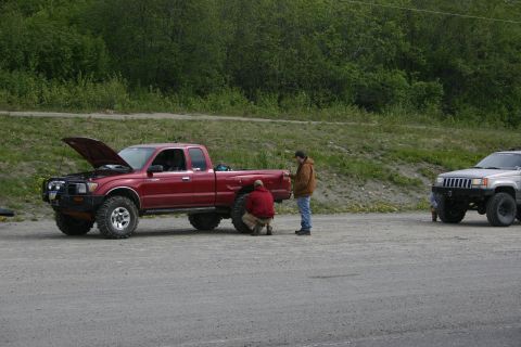 2003 Jeep TJ - Two individuals attend to a red pickup truck with its hood up, while a silver SUV is parked to the right.