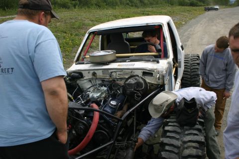 2003 Jeep TJ - Several people inspect a heavily modified pickup truck with oversized tires and an open engine bay on a dirt road.