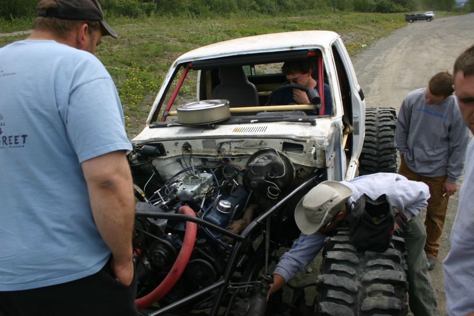 Modified pickup truck with large tires and exposed engine, people gathered around it