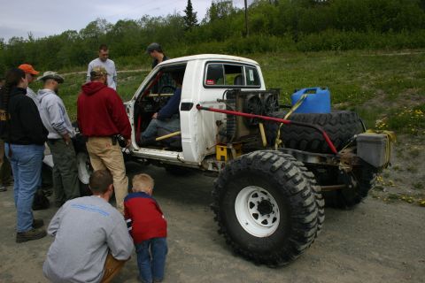 2003 Jeep TJ - A group of people gather around a heavily modified white truck with oversized tires and a roll cage.