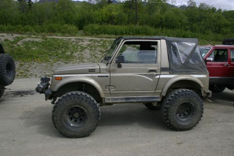 2003 Jeep TJ - A lifted Suzuki Samurai with aggressive off-road tires is parked on a dirt road, with a red SUV visible to its right.