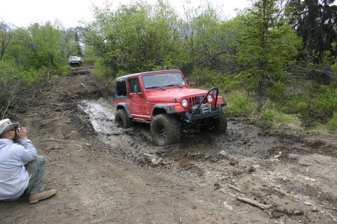 2003 Jeep TJ - A red Jeep Wrangler tackles a challenging muddy off-road course with another vehicle in the distance.