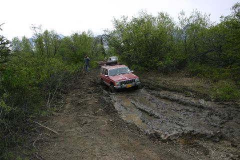 2003 Jeep TJ - A red Jeep Cherokee XJ struggles through a deep mud pit on an off-road trail.