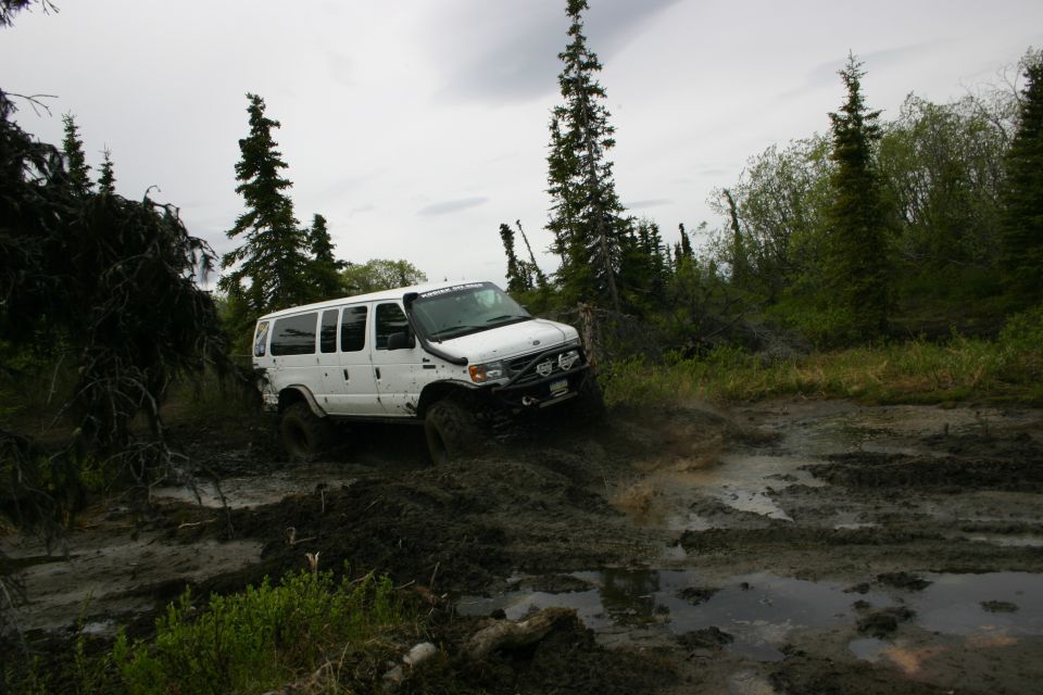 White van with snorkel and off-road tires driving through a muddy trail in a wooded area