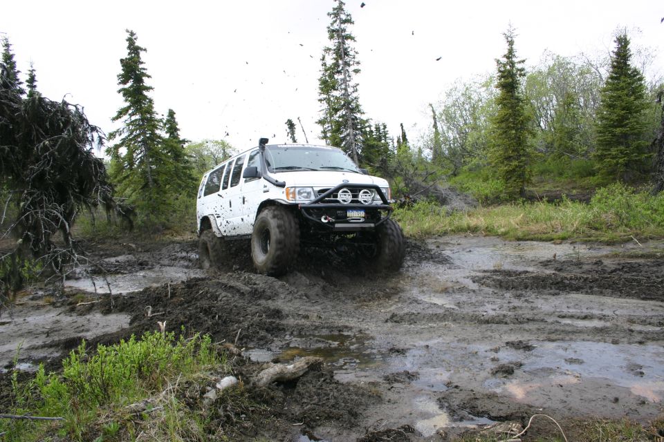 White SUV with snorkel and off-road bumper driving through a muddy trail with trees in the background