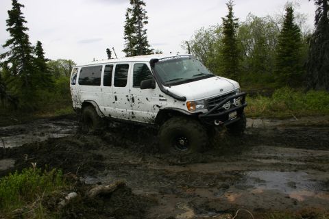 2003 Jeep TJ - A white van navigates a muddy off-road trail, kicking up mud as it goes.