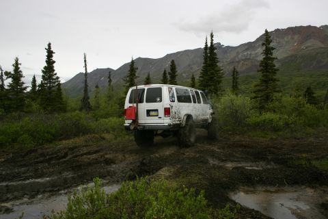 2003 Jeep TJ - A white van equipped for off-roading navigates a muddy path through a wooded area with mountains in the distance.