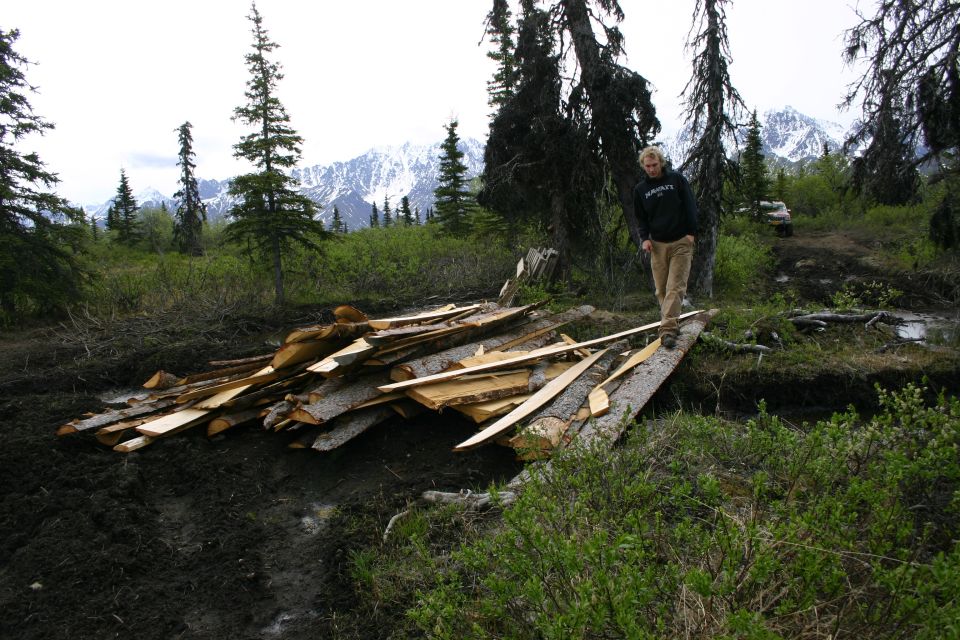 Person walks across a log and lumber bridge over a muddy area, with mountains in the background