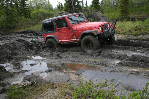 2003 Jeep TJ - A red 2003 Jeep TJ navigates a muddy trail, its tires deep in the muck.