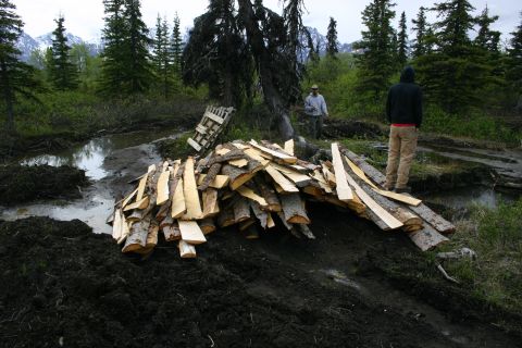 2003 Jeep TJ - Two individuals stand near a large pile of cut wood in a wet, natural landscape with trees and mountains visible.