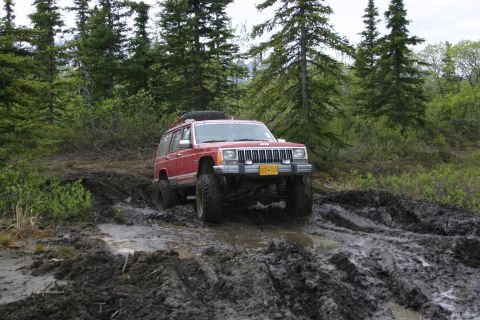 2003 Jeep TJ - A red Jeep Cherokee XJ navigates a challenging muddy off-road path surrounded by dense forest.
